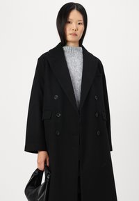 Svart dobbeltspent frakk laget av ull, med notched revers, store knapper og sidelommer; kombinert med en grå strikkegenser og svart veske.