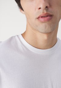 Close-up van het onderste deel van het gezicht en de nek van een jonge man die een eenvoudige witte T-shirt met ronde hals draagt tegen een neutrale achtergrond.