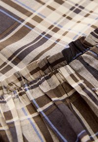 Tissu à carreaux dans des tons de beige, marron et bleu, présentant une texture froncée le long du bord et des largeurs de rayures variées dans un motif à carreaux.