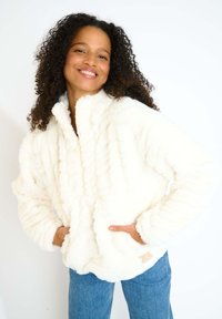 Giacca in felpa bianca e soffice con colletto alto, chiusura con zip e due tasche anteriori. Texture morbida e vestibilità rilassata. Jeans in denim azzurro chiaro.