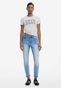 Camiseta gris claro con la impresión "LOVE DSGL", combinada con unos vaqueros ajustados azules y unos zapatos negros sin cordones. Diseño simple, look casual.