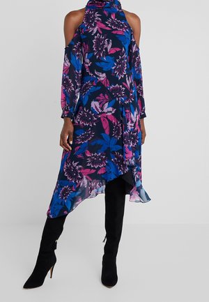 Femme portant une robe fleurie bleu marine à épaules dénudées avec des fleurs roses et bleues, associée à des bottes noires à talons montant jusqu'aux genoux.