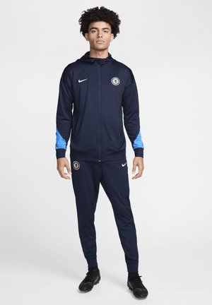 Ensemble de sport marine avec des accents bleu clair, comprenant une veste à capuche zippée et un pantalon assorti, fabriqué dans un tissu lisse et extensible.