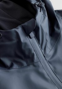 Gros plan sur une veste imperméable sombre avec une fermeture éclair à l'avant, des coutures surpiquées et une capuche doublée en mesh avec des cordons de serrage réglables.
