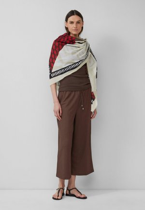 Châle tissé avec un motif géométrique rouge et noir sur un fond ivoire, associé à un haut côtelé marron et à un pantalon large court.