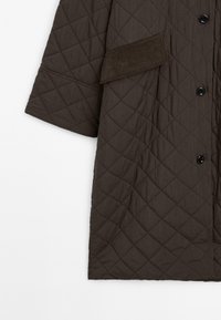 Gestepptes braunes Kleidungsstück mit weiten Ärmeln, ausgestattet mit schwarzen Knöpfen und einer Kordstoff-Akzenttasche auf der linken Seite. Glatte Textur, Rautenmuster.