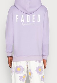 Felpa con cappuccio lavanda con la scritta "FADED" bianca sulla schiena, caratterizzata da una texture morbida e spalle scese. Abbinata a pantaloni con motivo floreale.