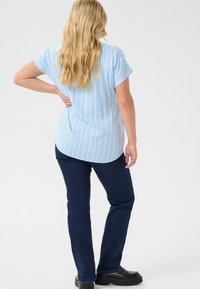 Camisa azul clara con rayas blancas verticales, de mangas cortas y con dobladillo curvado, combinada con vaqueros oscuros y zapatos negros. Vista trasera.