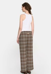 Kobieta z kręconymi włosami, nosząca duże kolczyki w kształcie kół, białą bluzkę bez rękawów, brązowe, w kratę szerokie spodnie oraz brązowe buty, stoi tyłem.