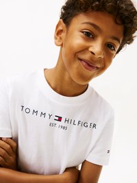 Λευκό βαμβακερό μπλουζάκι με εκτύπωση "TOMMY HILFIGER EST. 1985" σε μαύρο χρώμα, με κόκκινη και μπλε λογότυπο λεπτομέρεια στο μανίκι. Κοντινό πλάνο παιδιού.