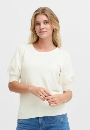 Light cream short-sleeve sweater with puffed sleeves, ribbed texture, and crew neck. Paired with blue denim jeans.
