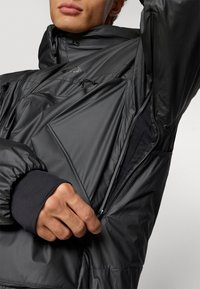 Tmavo šedá zateplená bunda s vysokým golierom, bočnými vreckami na zips a elastickými manžetami. Hladká tkanina s elegantným a funkčným dizajnom.