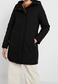 Abrigo negro con capucha que presenta un cierre frontal con cremallera, dos bolsillos laterales y un exterior liso. El diseño incluye un ajuste holgado y costuras visibles.