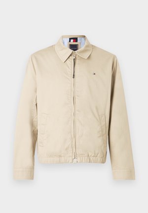 Beige jakke med krave, frontlynlås og to sidelommer. Har et stribet inderfor og et lille logo på brystet.