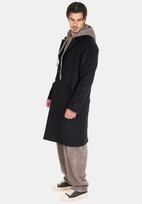 Uomo in soprabito nero sovrapposto a un hoodie marrone, pantaloni marroni larghi e scarpe da ginnastica nere, in piedi e che guarda a sinistra su uno sfondo bianco.