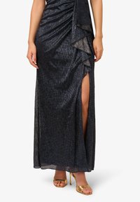 Dlouhá, tmavě modrá večerní šaty s plisovaným designem a vrstvenými akcenty, s vysokým rozparkem a spárované se zlatými sandály na podpatku.