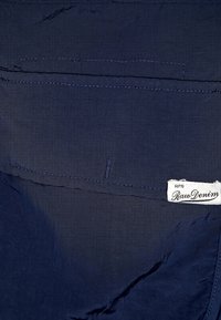 Navy fabric with a subtle textured pattern, featuring a pocket and a sewn label that reads "Raw Denim" with a numerical code.