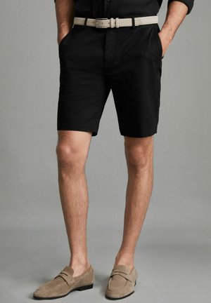 Hombre con pantalones cortos negros, cinturón beige y mocasines de ante marrón claro, de pie con las manos en los bolsillos contra un fondo gris.