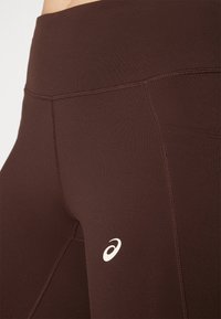 Leggings atléticos marrones con una cinturilla alta, textura suave, costuras reforzadas y un pequeño logo blanco en el lado izquierdo.