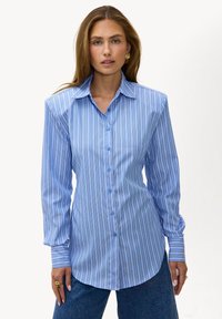 Femme portant une chemise à rayures verticales bleu et blanc à boutons, manches longues, et un jean bleu, debout devant un fond blanc.