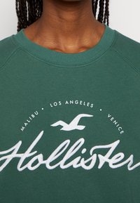 Vihreä collegepaita, jossa on resorinen kaulus ja valkoinen kirjailtu teksti "Hollister" sekä pieniä grafiikoita ja yläpuolella kaupunkien nimiä.