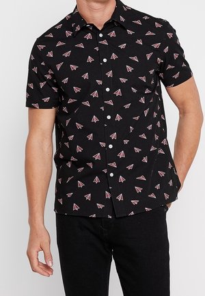 Schwarzes kurzärmeliges Hemd mit Knöpfen und einem Muster aus rosa-weißen Papierfliegern, getragen von einem Mann mit einer Hand in der Tasche.