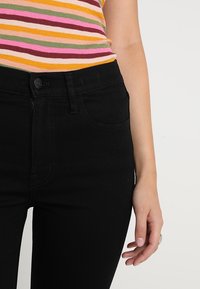 Svarta denimjeans med hög midja, med knappstängning fram, standardfickor och en slät textur. Modell bär en randig topp.