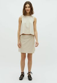 Beige ärmelloses Oberteil mit einem cropped Design, kombiniert mit einem passenden Wickelrock. Strukturierter Stoff, minimale Nähte und eine seitliche Tasche. Schwarze Schnürschuhe.