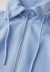 Felpa con zip di colore azzurro chiaro realizzata in tessuto texturizzato, dotata di un colletto alto, zip argento e lacci con estremità in metallo.