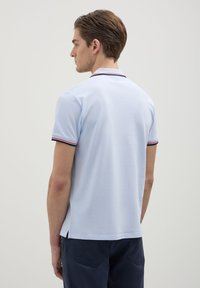 Giovane uomo di spalle che indossa una polo azzurro chiaro con colletto e maniche rigate in navy e rosso, e pantaloni blu scuro.
