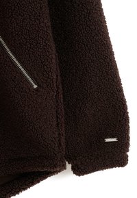 Bruine fleec Jas met een ritszak, textuur op het oppervlak en een metalen merketiket op de onderrand. Zacht en comfortabel materiaal.