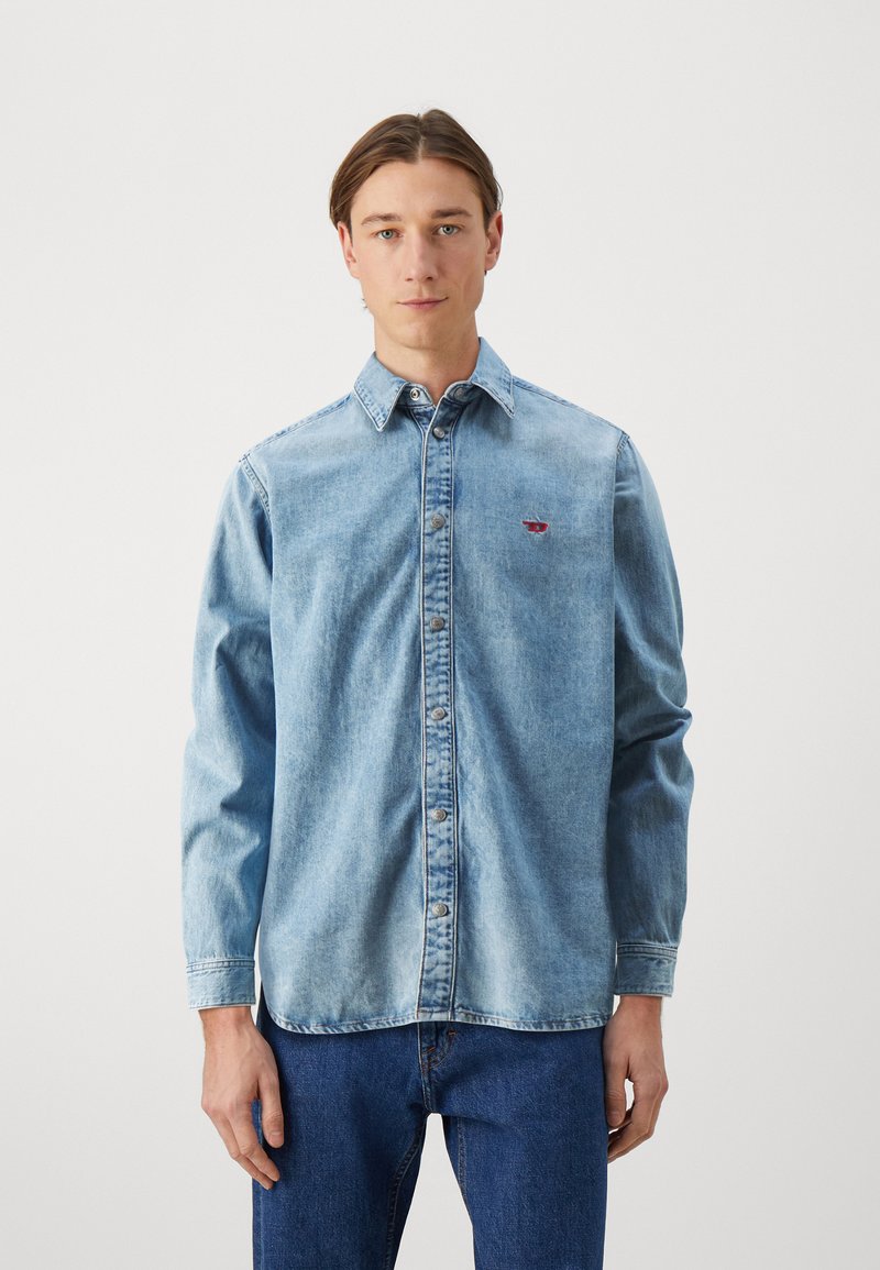 Light blue denim shirt with long sleeves, button-up front, and a small red logo on the left chest, featuring a classic collar and relaxed fit.