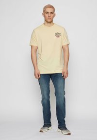 Noor blondide lühikeste juustega noormees, kes kannab heledat kollast "Brunch Sundays" t-särki, siniseid teksaseid ja halli spordijalatsit, seistes ühetaolise tausta vastas.