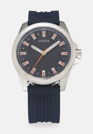 Montre en acier inoxydable argenté avec un bracelet en caoutchouc bleu foncé. Présente un cadran foncé avec des index horaires argentés et rouges, et une aiguille des secondes orange.