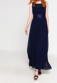 Kvinna klädd i en ärmlös aftonklänning i marinblått med ett satinbälte och svarta remsandaler med hög klack, går framåt.