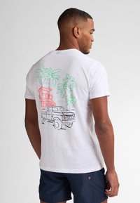 T-shirt bianco in cotone con una grafica colorata di un camion, una torre di salvataggio e palme sul retro. Maniche corte, colletto girocollo.