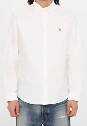 Homme portant une chemise blanche à manches longues avec un petit logo brodé sur la poitrine et un jean bleu.