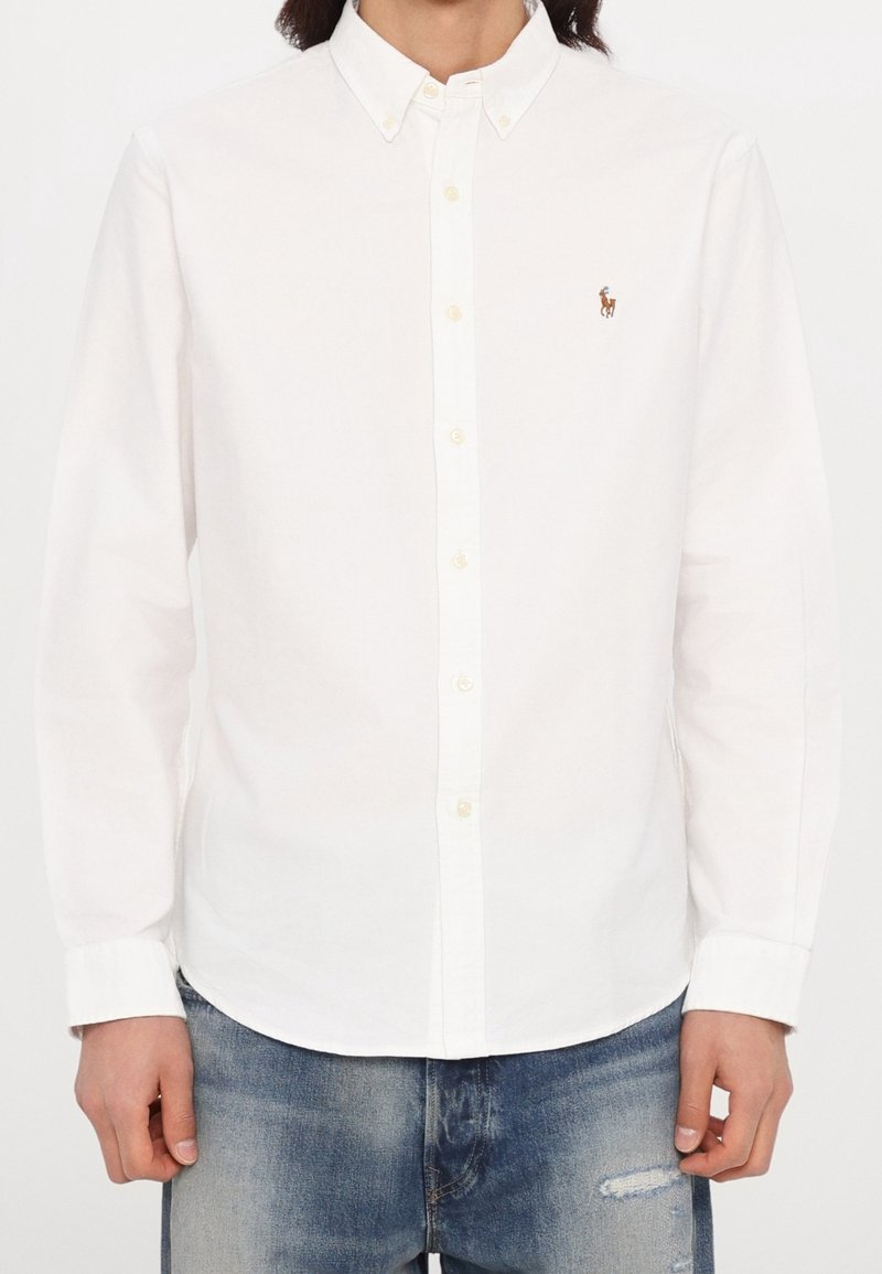 Homme portant une chemise blanche à manches longues avec un petit logo brodé sur la poitrine et un jean bleu.