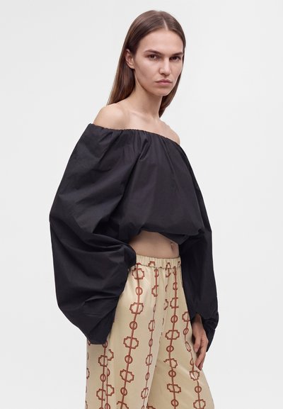 Jeune femme aux longs cheveux porte un haut noir à épaules dénudées avec des manches volumineuses et un pantalon beige avec un motif géométrique rouge.