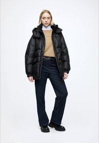 Crna jakna s kapuljačom, s prešanim dizajnom i sjajnim završetkom, nošena preko bež džempera i tamnoplavih traperica.