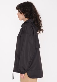 Veste à capuche noire en matériau léger, dotée d'une poche avant et de poignets élastiques. Design ample avec un ourlet descendant.