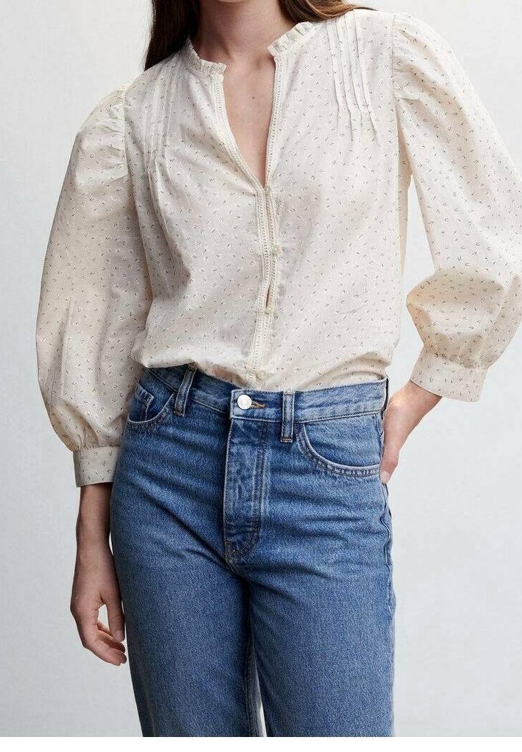 Femme portant un chemisier long crème texturé avec des manches bouffantes et un jean bleu taille haute, main reposant sur la hanche.