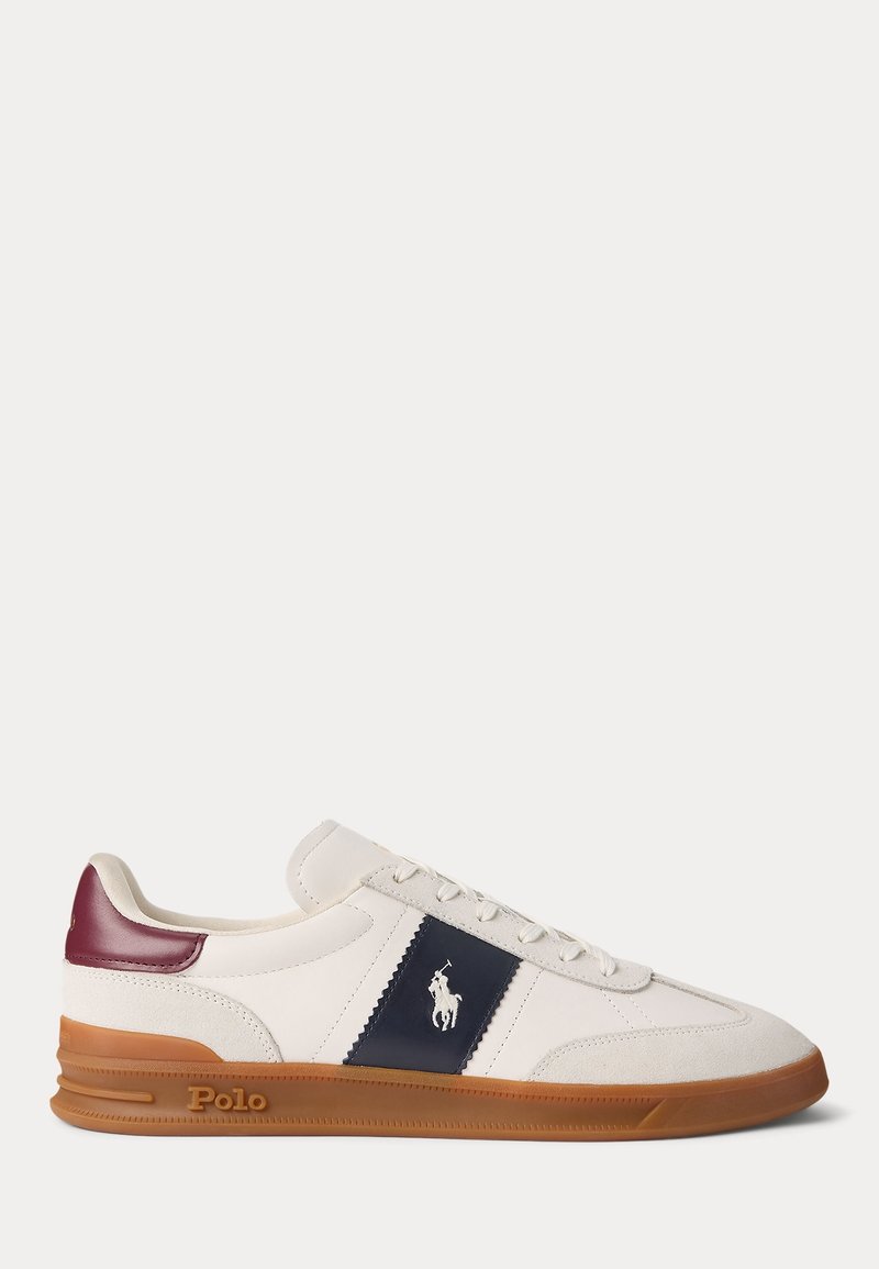 Zapatilla de cuero blanca con una franja azul marino, detalle en burdeos en el talón y suela de goma color marrón. Presenta cordones y un pequeño logo en el costado.