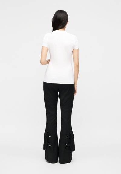 Γυναίκα στέκεται με την πλάτη στην κάμερα, φοράει λευκό κοντομάνικο πουκάμισο και μαύρο παντελόνι με φαρδιά πόδια, πλαϊνές τσέπες και λεπτομέρειες με αγκράφες.