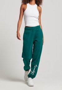 Türkise Jogginghose mit lockerer Passform, elastischem Bund und weißem, kursive Logo am linken Bein. Getragen mit einem weißen Tanktop und weißen Schuhen.
