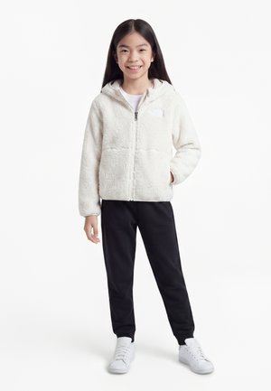 Lachend meisje staand, gekleed in een witte fleecejas met ritssluiting, zwarte broek en witte sneakers tegen een effen witte achtergrond.