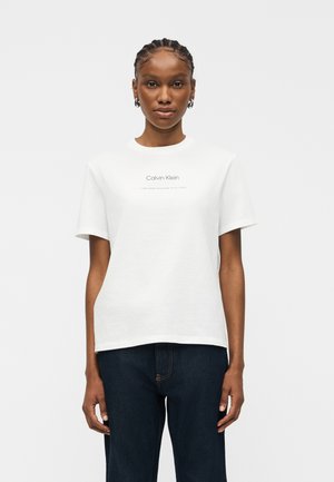 Nő fonott hajjal, fehér Calvin Klein pólót és sötétkék farmert viselve, sima világos háttér előtt áll.