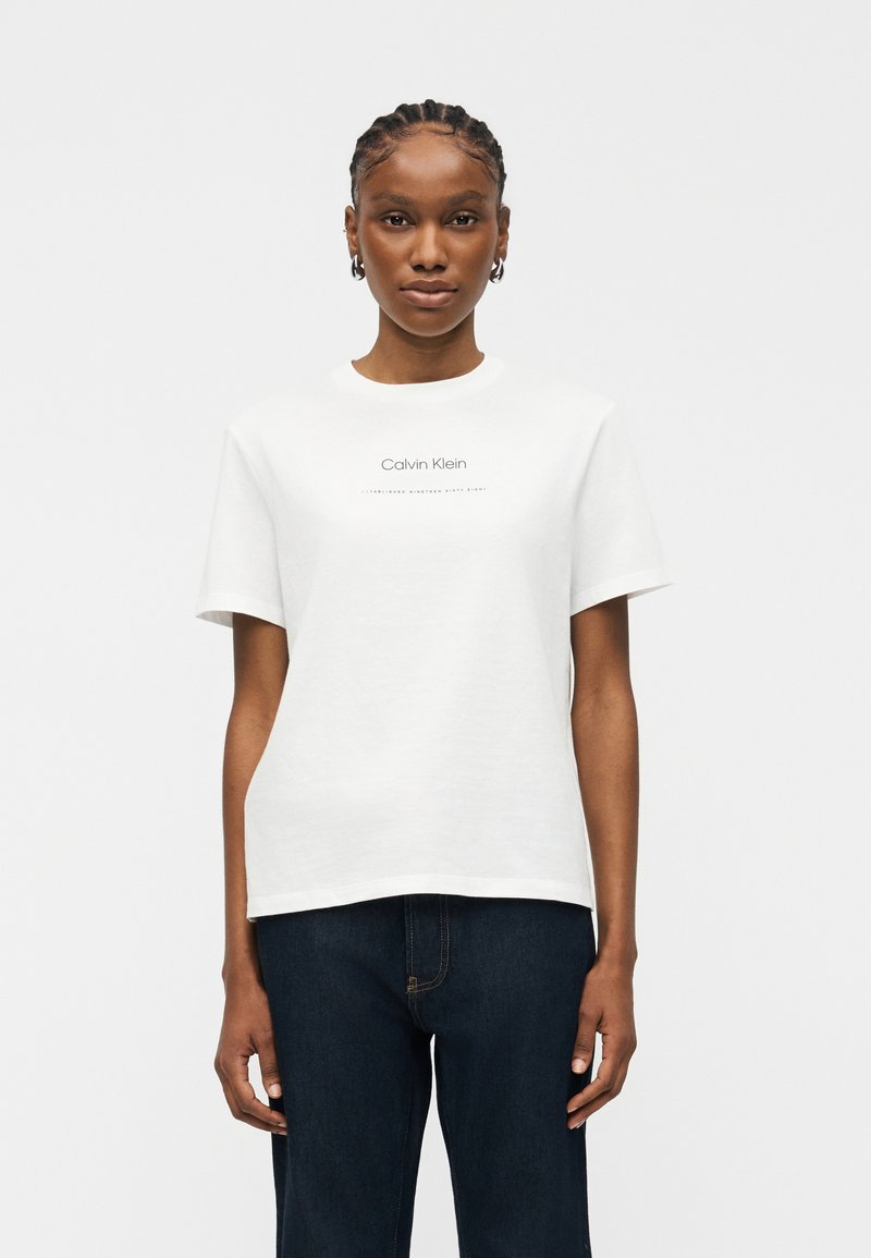 Γυναίκα με πλεγμένα μαλλιά που φοράει λευκό μπλουζάκι Calvin Klein και σκούρο μπλε τζιν, στέκεται μπροστά σε απλό ανοιχτό φόντο.