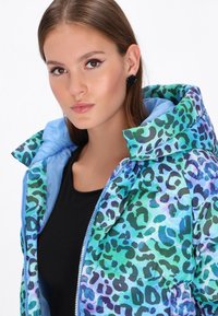 Daunenjacke mit einem lebhaften grünen und blauen Leopardenmuster, ausgestattet mit einem hellblauen Innenfutter und einer großen Kapuze. Reißverschluss vorne.