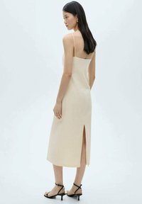 Ærmeløs midi-kjole i off-white stof med en lige silhuet, der har en sideslids og minimale stropper. Båret med sorte strophæle.